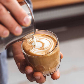 Hand gießt Milchschaum in Kaffeeglas mit Latte-Art-Motiv