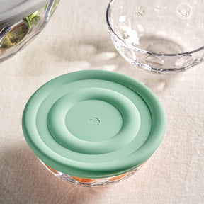 Silikondeckel CUCINA auf Glas, daneben Wasserglas und Ölkaraffe auf Tuch