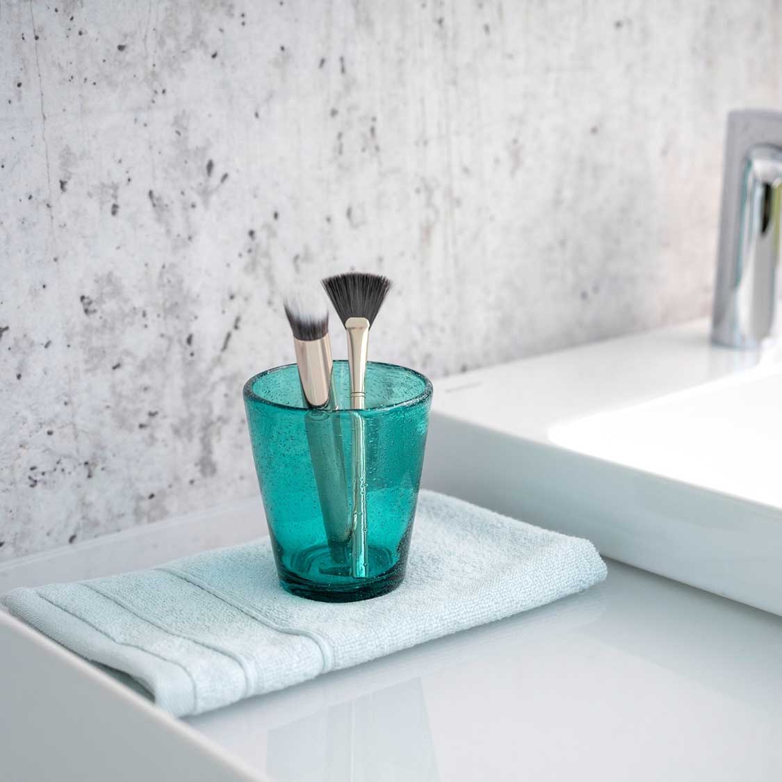Türkises Glas mit Pinseln auf Handtuch im Badezimmer