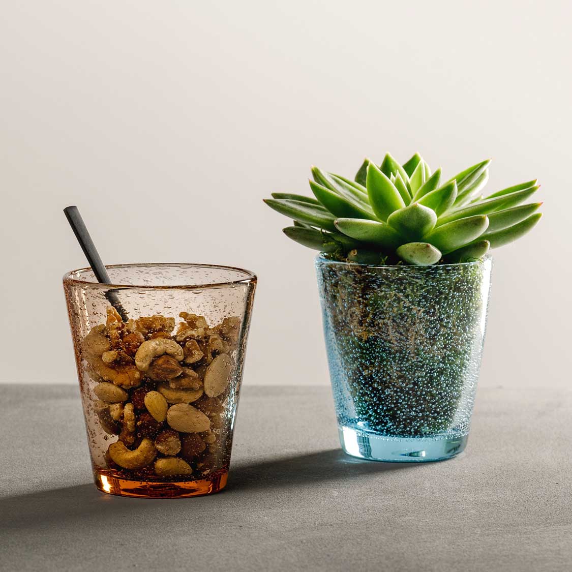 Trinkglas mit Cola und Cashewnüssen neben Sukkulente auf grauem Untergrund