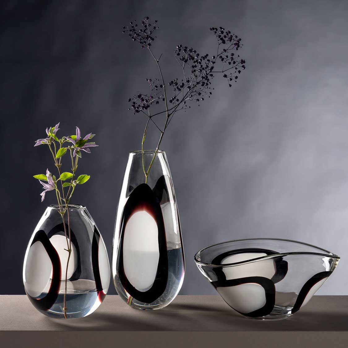 Vase OCCHI BIANCHI mit schwarzen Augenmustern, gefüllt mit getrockneten Blüten, neben weiteren Glasobjekten der Serie