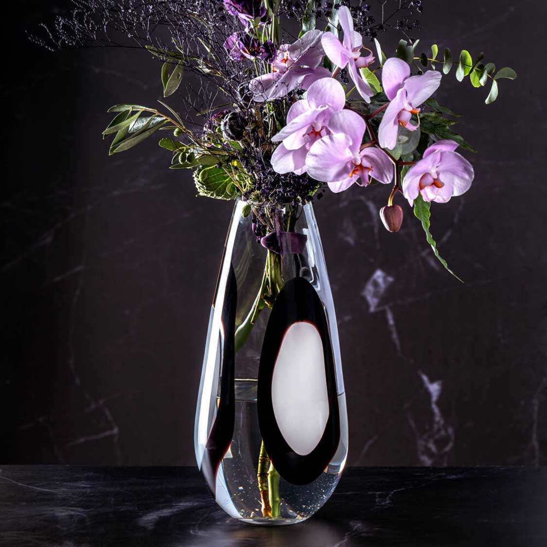 Vase OCCHI BIANCHI mit lila Orchideen und Grünpflanzen auf dunklem Hintergrund