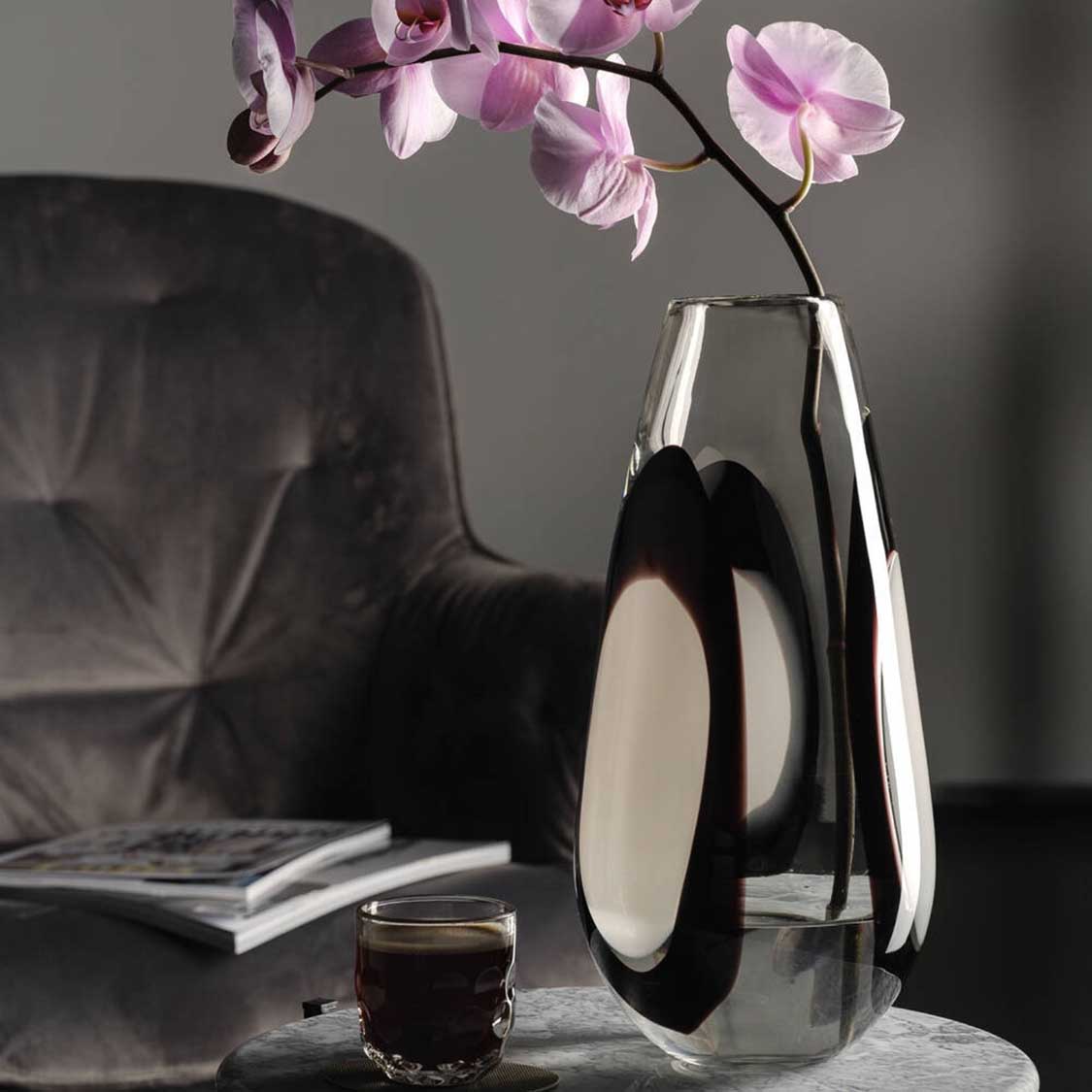 Vase OCCHI BIANCHI mit Orchideenzweig auf Marmortisch, neben Glas und Stuhl