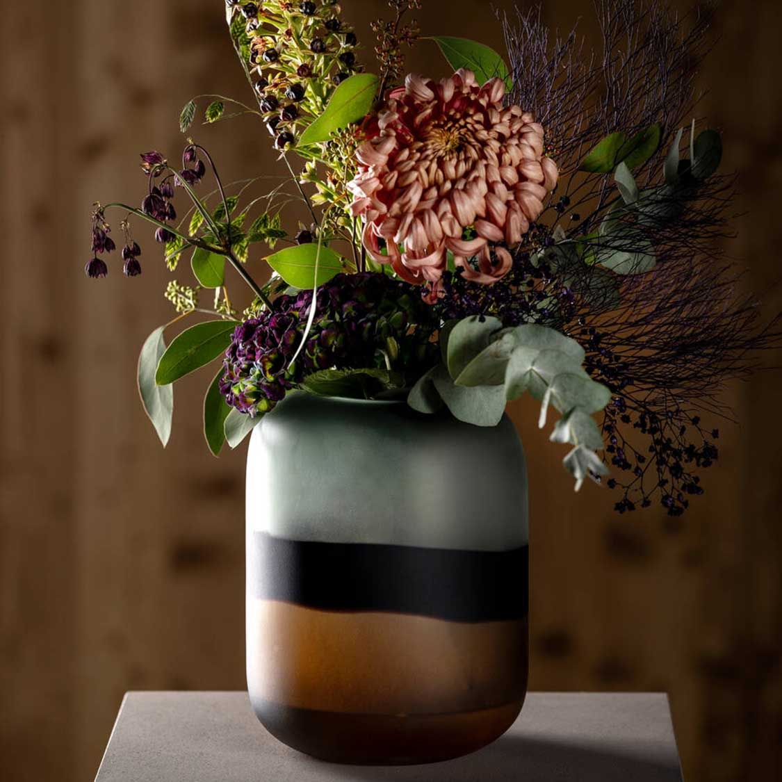 Vase CORSO SABBIA mit Schnittblumen und Grünpflanzen gefüllt, auf grauem Untergrund vor braunem Hintergrund