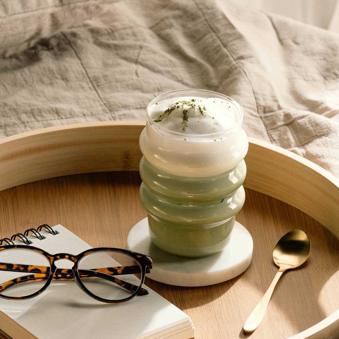 Trinkglas SOLE mit Matcha-Latte auf Holztablett, Brille und Löffel