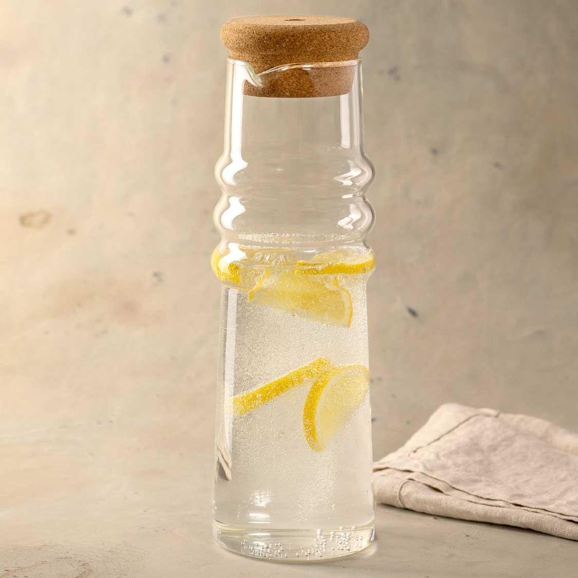 Karaffe SOLE mit Korkstopfen, gefüllt mit Wasser und Zitronenscheiben, auf hellem Untergrund