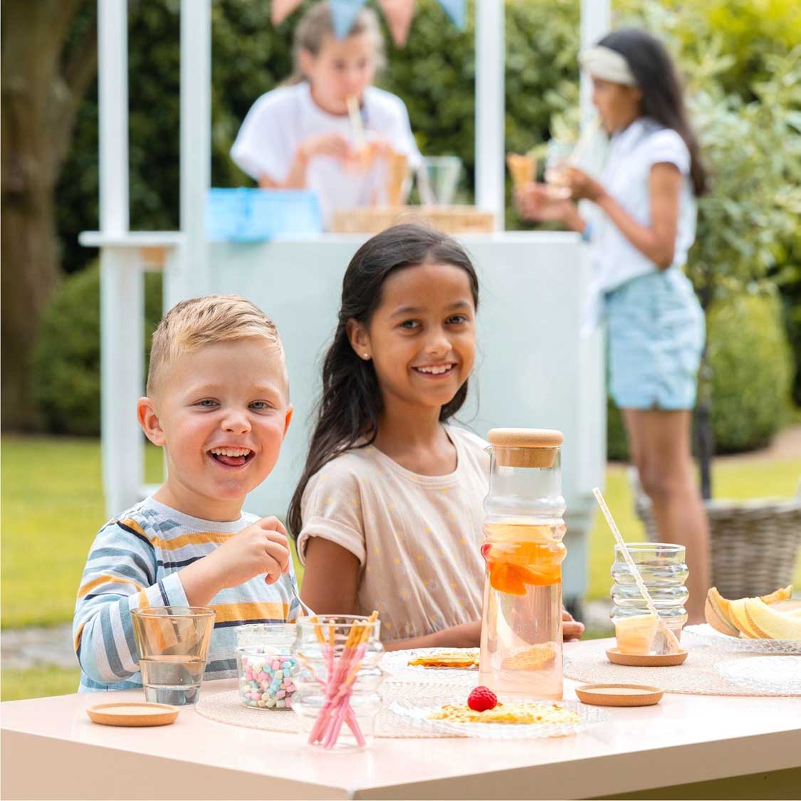 Karaffe SOLE mit Korkstopfen auf Gartentisch, Kinder beim Getränkeausschank