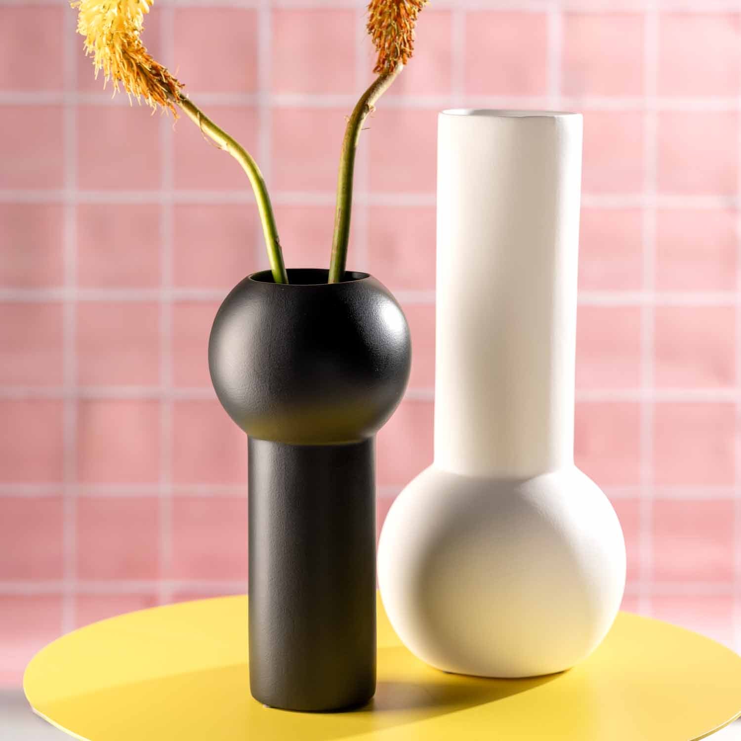 Schwarze Keramikvase BRERA mit getrockneten Blüten, daneben weiße Vase auf gelbem Untergrund vor rosa Fliesenhintergrund