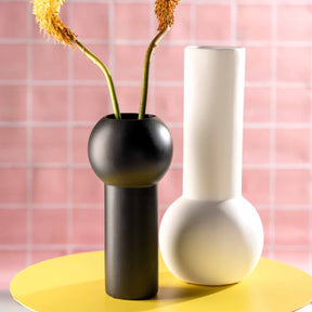Schwarze Keramikvase BRERA mit getrockneten Blüten, daneben weiße Vase auf gelbem Untergrund vor rosa Fliesenhintergrund