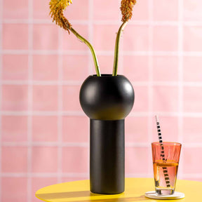 Keramikvase BRERA schwarz mit Trockenblumen, neben Glas mit Getränk auf gelbem Tisch