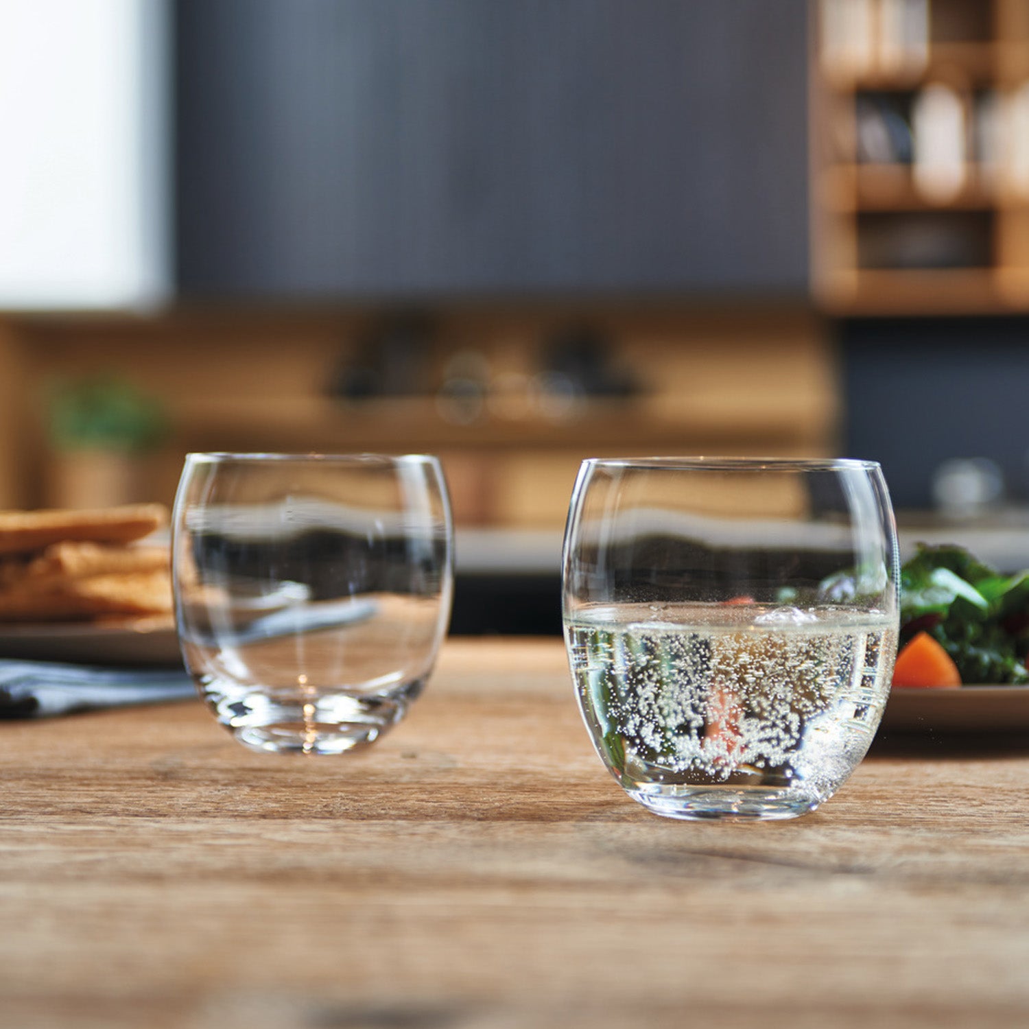 Zwei Trinkgläser CHEERS auf Holztisch in Küchenumgebung, eines mit Wasser gefüllt