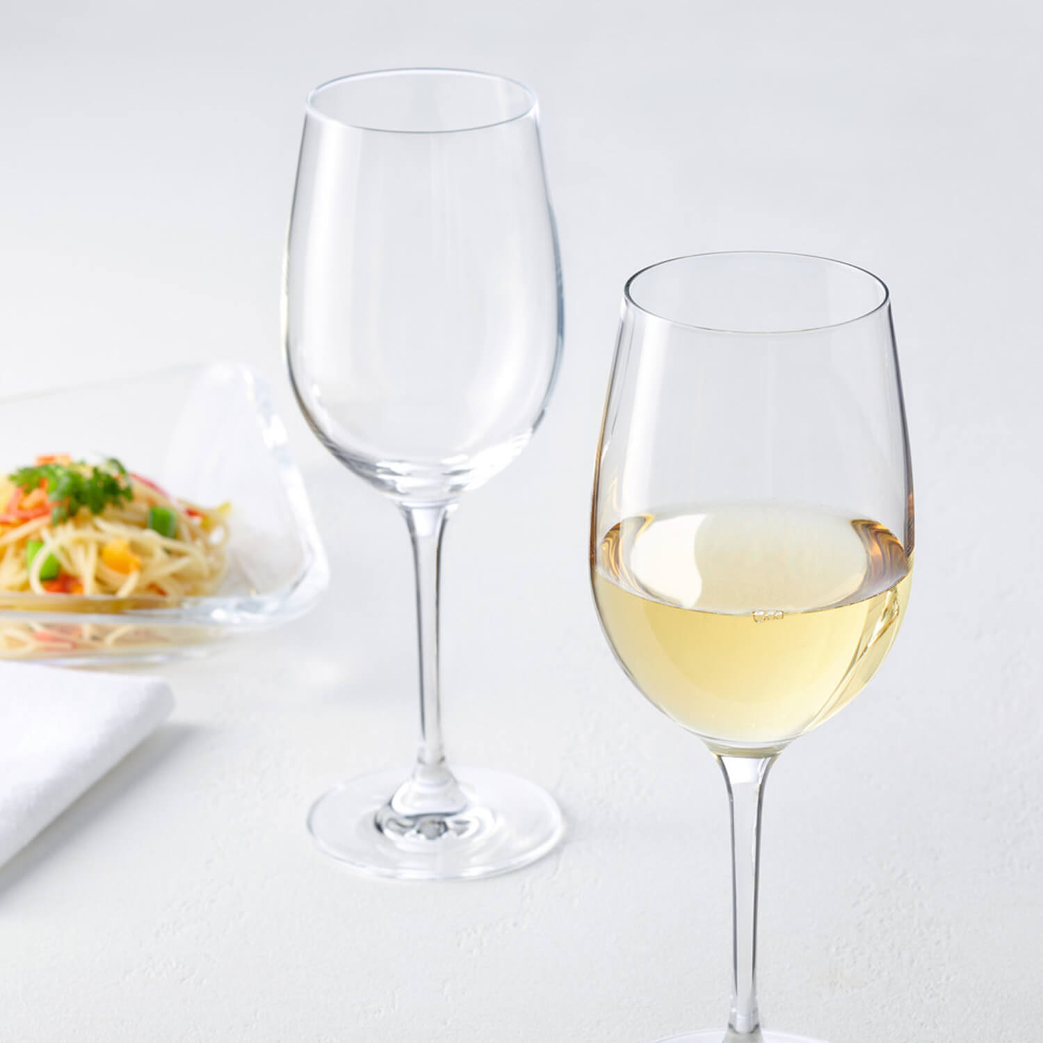 Weißweinglas CIAO+ mit Wein gefüllt neben leerem Glas, Teller mit Pasta im Hintergrund