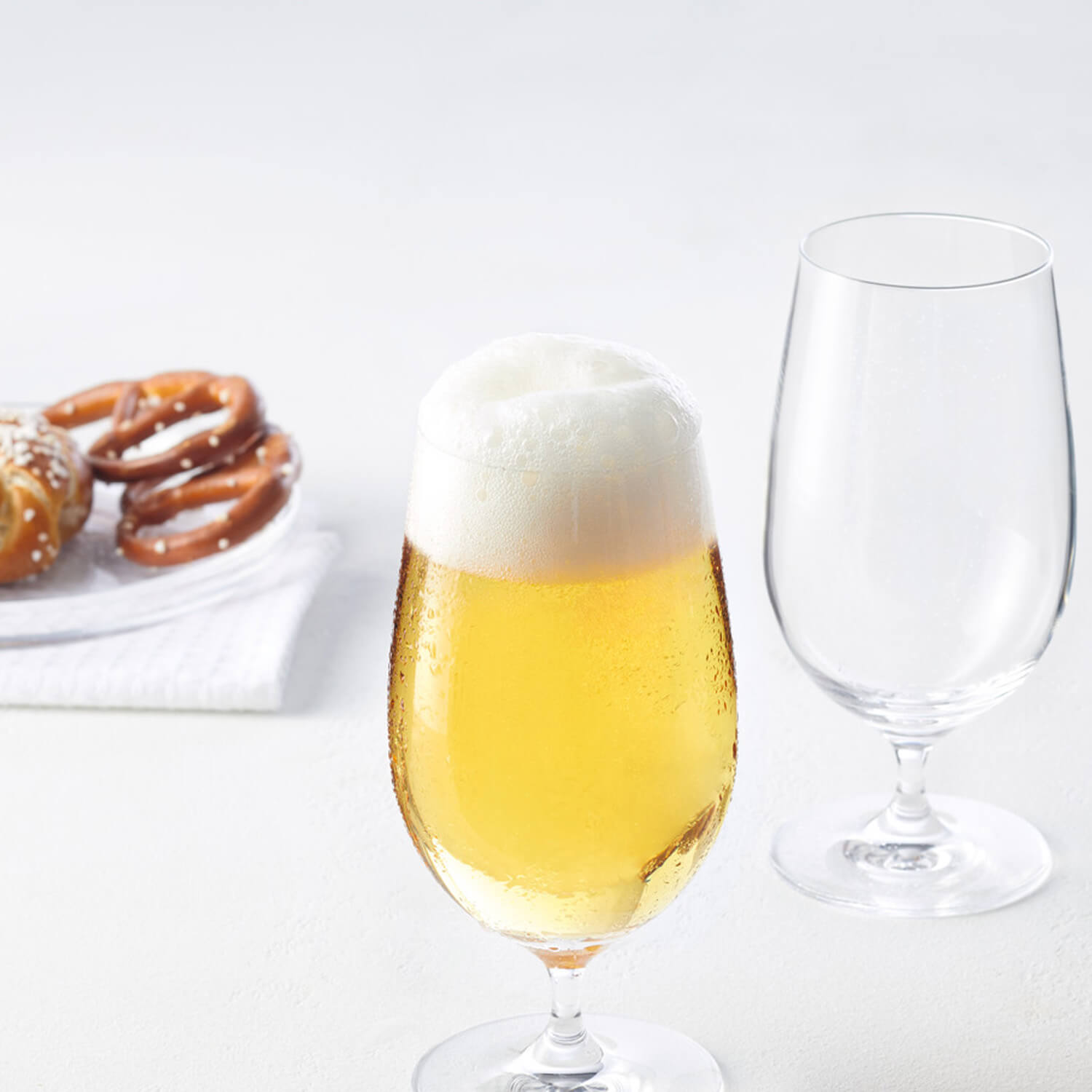 Bierglas CIAO+ mit Bier und Schaum, leeres Glas daneben, Brezel im Hintergrund