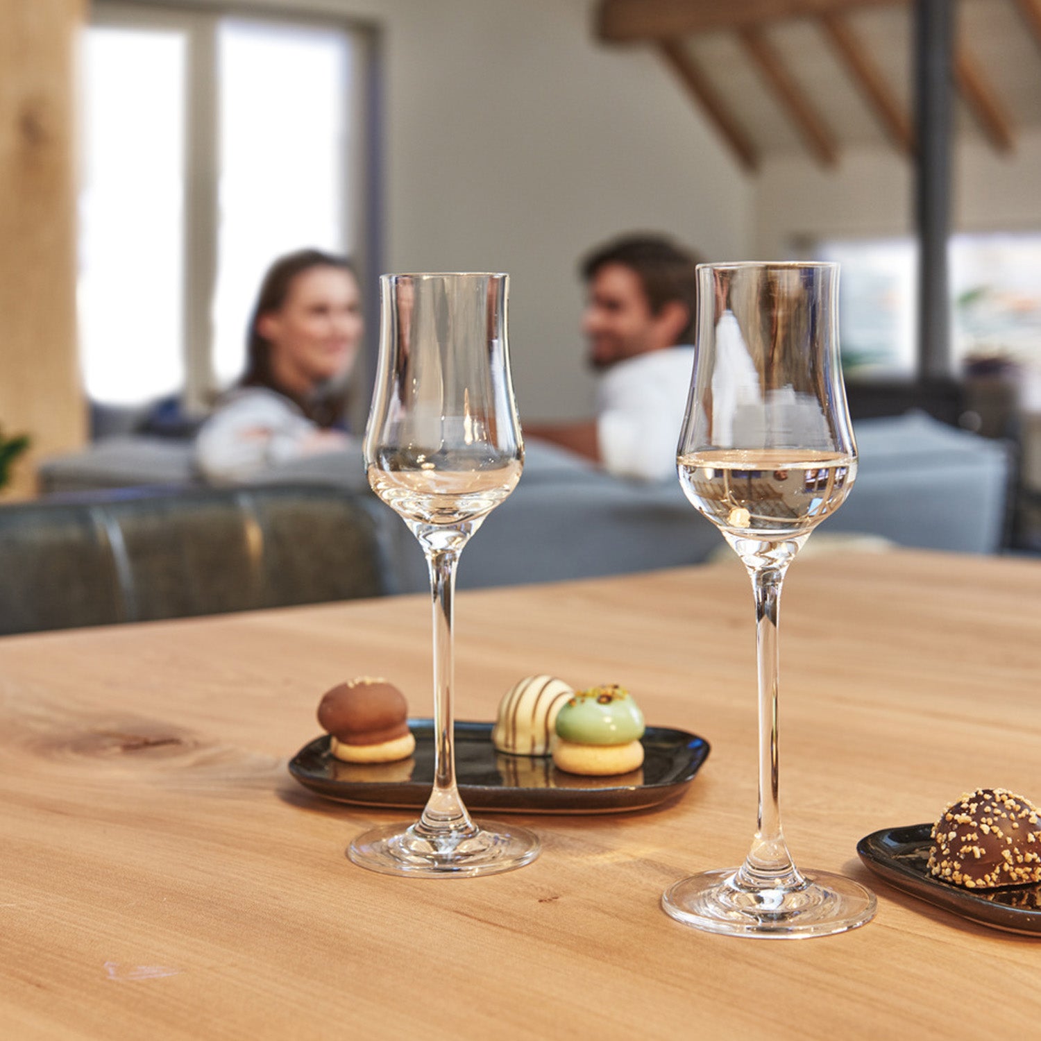 Zwei Grappagläser CIAO+ auf Holztisch mit Gebäck, Personen im Hintergrund auf Sofa
