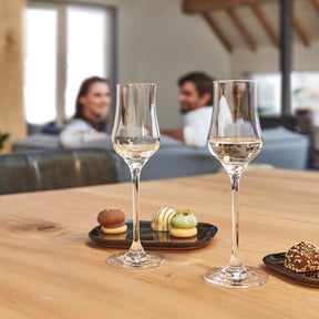 Zwei Grappagläser CIAO+ auf Holztisch mit Gebäck, Personen im Hintergrund auf Sofa
