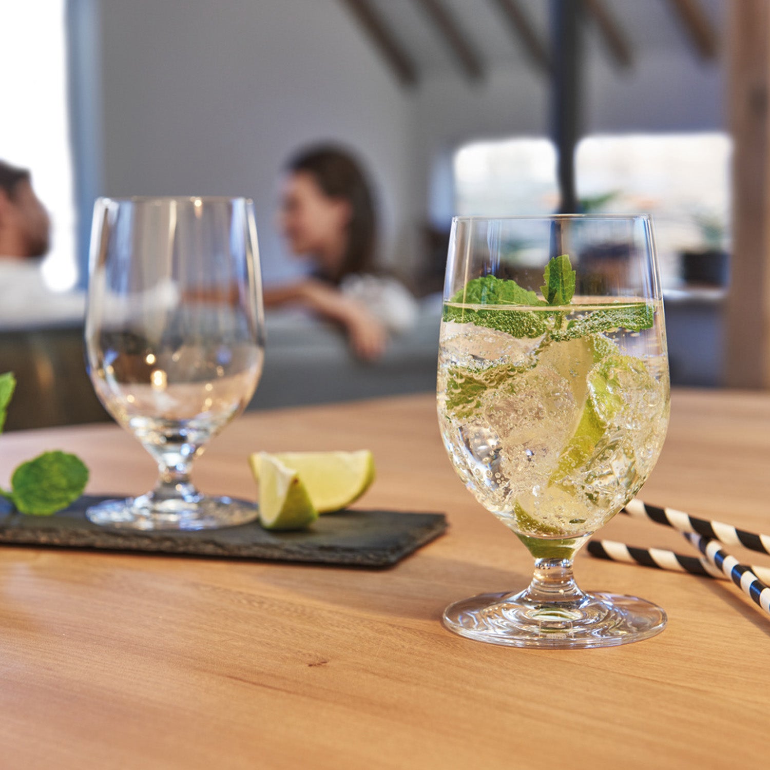 Wasserglas CIAO+ mit Minze und Limette auf gedecktem Tisch, Menschen im Hintergrund