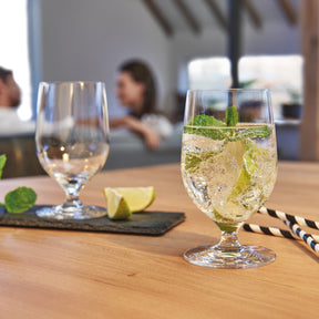 Wasserglas CIAO+ mit Minze und Limette auf gedecktem Tisch, Menschen im Hintergrund