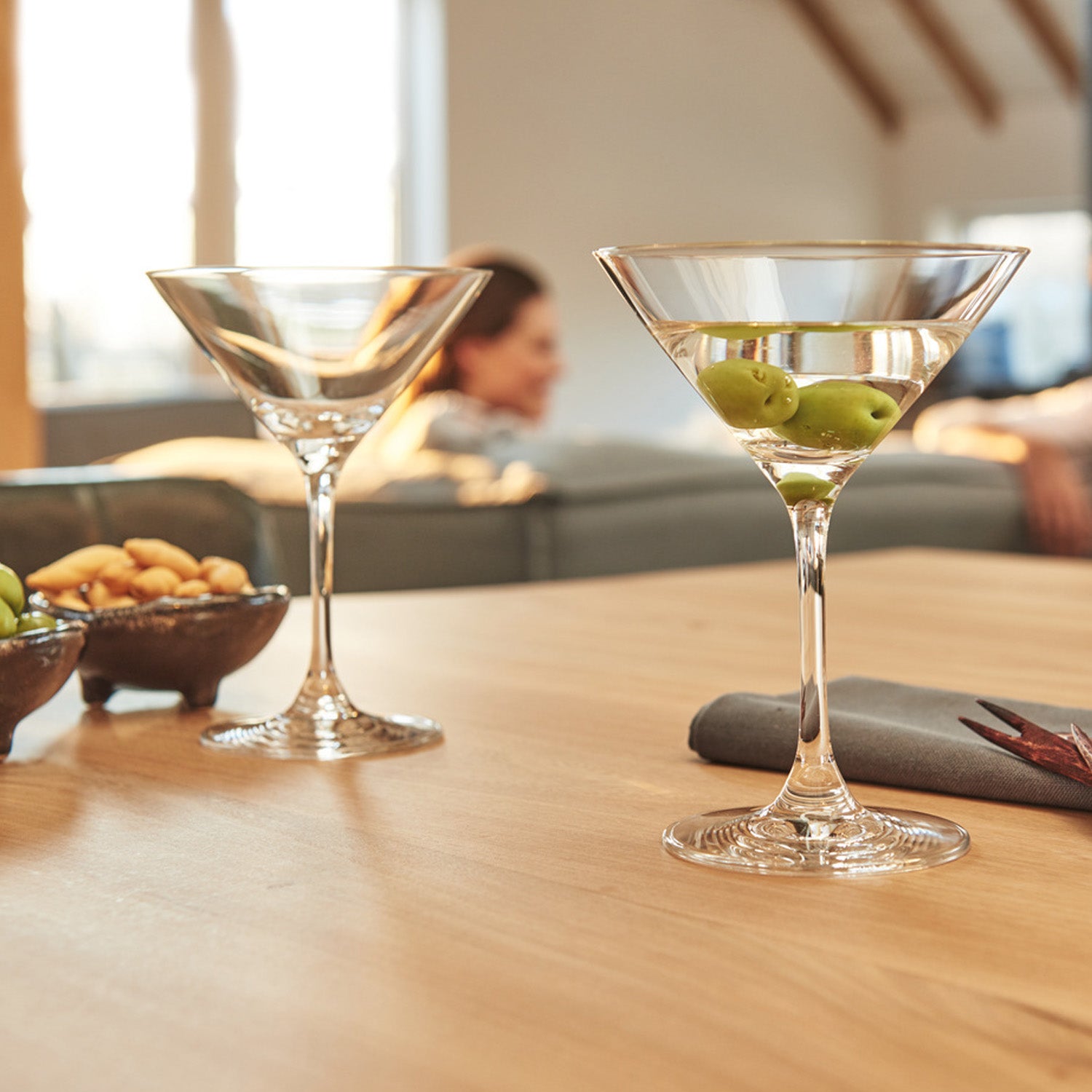 Cocktailgläser CIAO+ auf Holztisch mit Oliven, im Wohnzimmer mit Personen im Hintergrund
