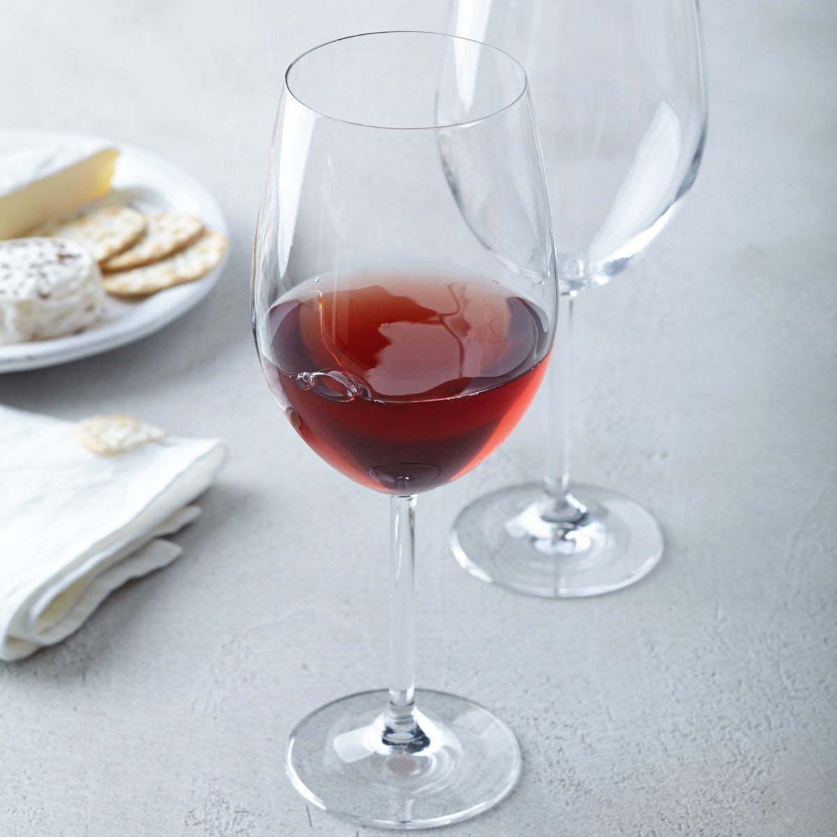 Bordeauxglas DAILY gefüllt mit Rotwein, daneben leeres Glas, gedeckter Tisch mit Käse und Crackern