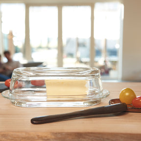Butterdose CIAO aus Glas mit Butter auf Holztisch, Buttermesser daneben, helles Tageslicht von Fensterfront