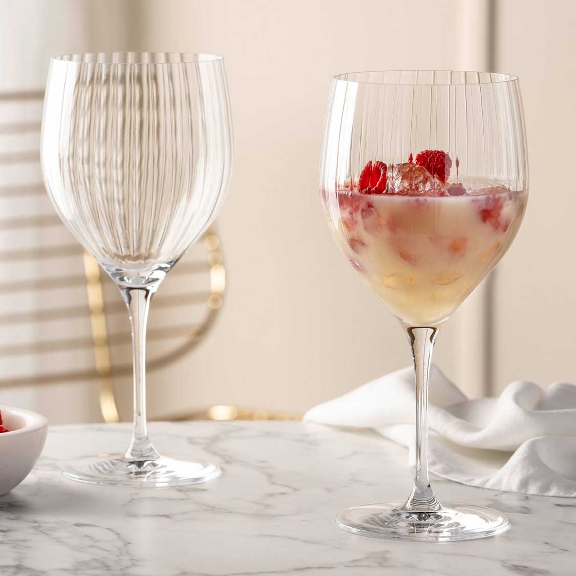 Zwei Cocktailgläser POESIA auf Marmortisch, eines leer, eines mit Getränk und Himbeeren gefüllt