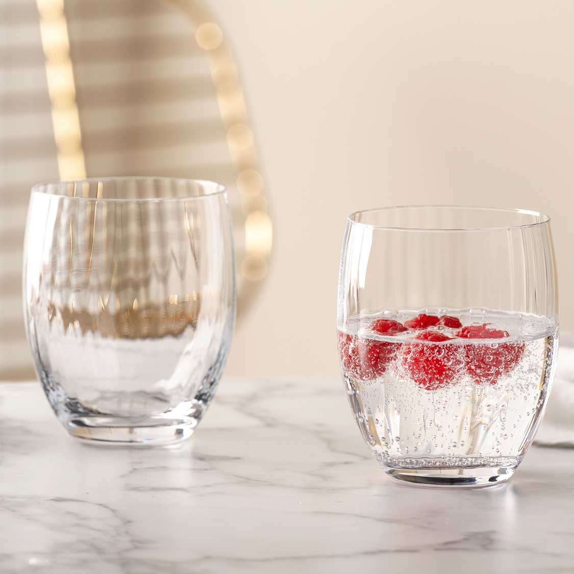 Zwei Trinkgläser POESIA auf Marmortisch, eines leer, eines mit Wasser und roten Beeren gefüllt