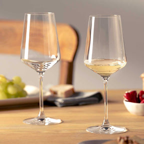 Zwei Rieslinggläser PUCCINI auf Holztisch, eines leer, eines mit Weißwein gefüllt, Obst und Schüssel im Hintergrund
