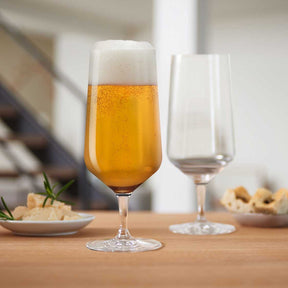 Bierglas PUCCINI mit Bier gefüllt auf Holztisch, daneben leeres Glas und Nüsse