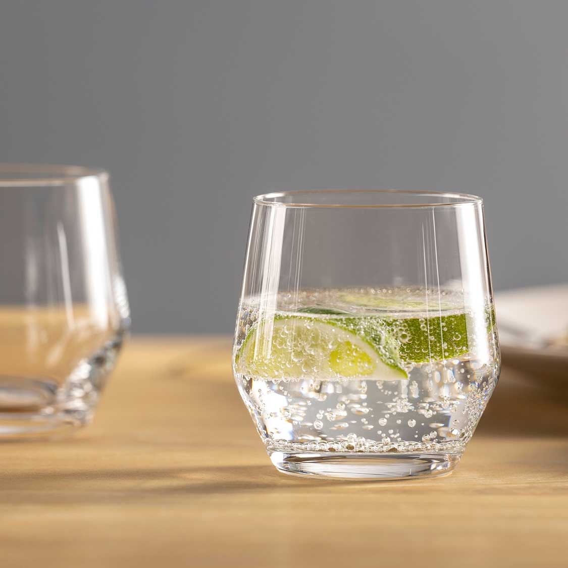 Trinkglas PUCCINI mit Limette und Eiswürfeln auf Holztisch, weitere Gläser im Hintergrund