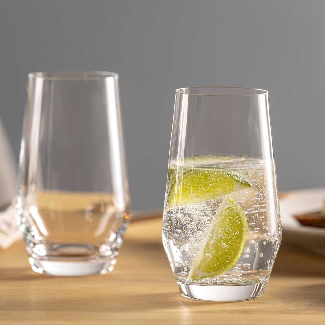 Zwei Trinkgläser PUCCINI auf Holztisch, eines leer, eines mit Wasser, Limette und Eiswürfeln gefüllt