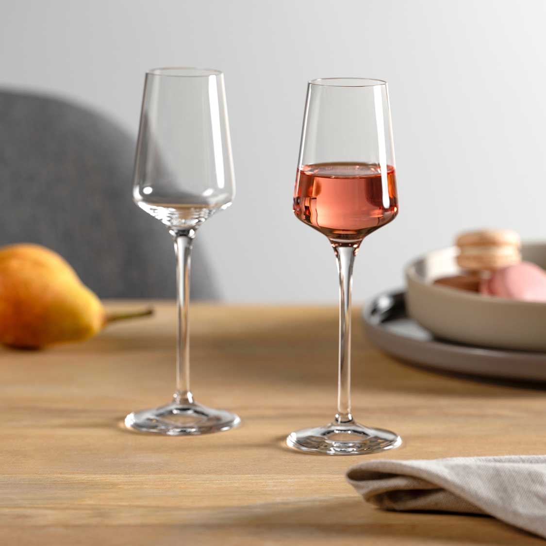 Zwei Grappagläser PUCCINI auf Holztisch, eines leer, eines mit rosé Flüssigkeit gefüllt, Tischgedeck im Hintergrund