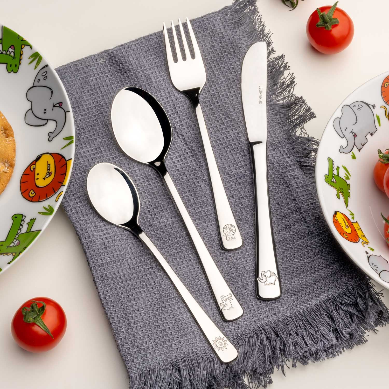Kinderbesteck BAMBINI 4-teilig silber auf grauem Tuch, arrangiert mit Tellern und Tomaten
