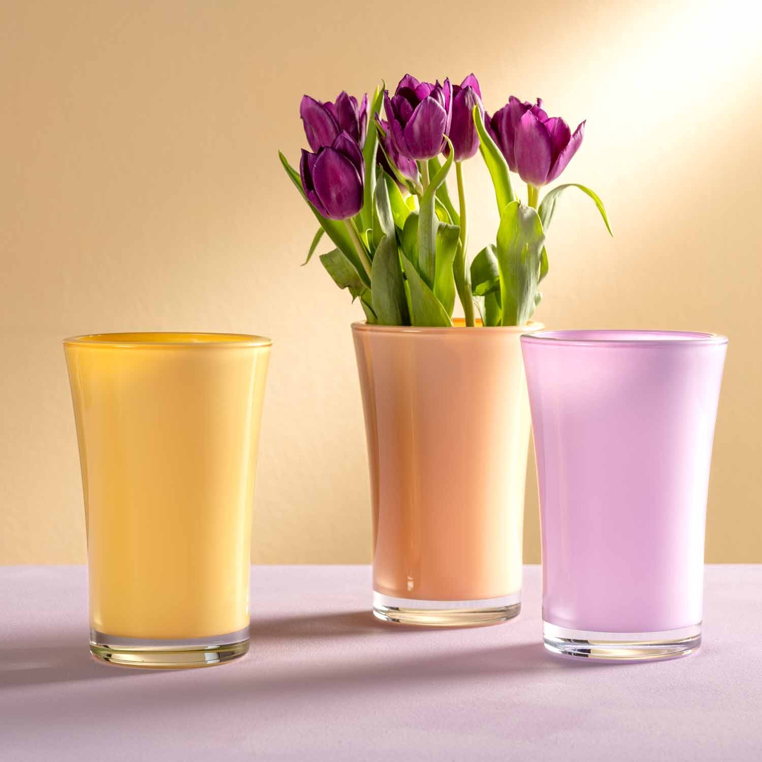 Drei Gläser mit gelber, pfirsichfarbener und lila Flüssigkeit, mittleres Glas mit lila Tulpen