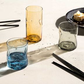 Trinkglas VESUVIO in Amber und weiteren Farben auf weißer Oberfläche mit Essstäbchen und Schale