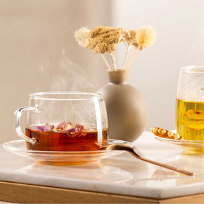 Teetasse mit rotem Tee, daneben Glas mit gelblichem Getränk, getrocknete Blumen in Vase, Teeblätter auf Untergrund