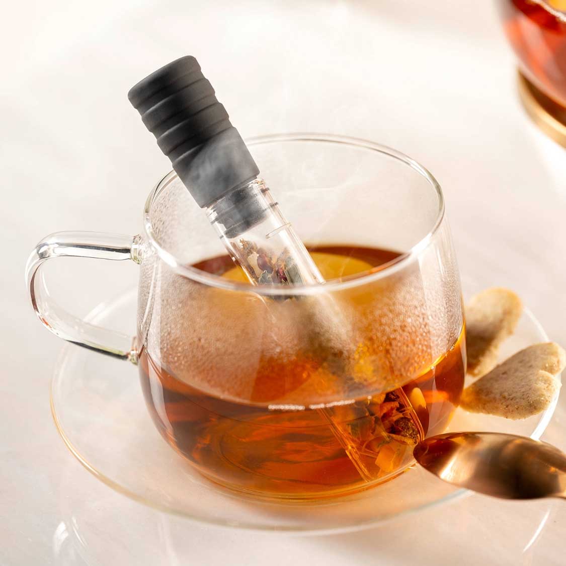 Teeinfuser in Glastasse mit heißem Tee, daneben Teelöffel auf Untertasse