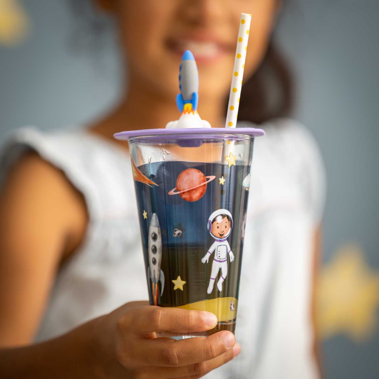 Kind hält Trinkbecher mit Raketen- und Astronauten-Motiv und lila Deckel