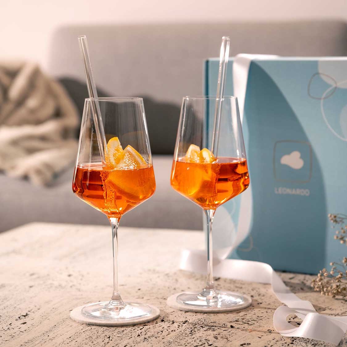 Zwei Weißweingläser PUCCINI mit Orangensaft und Eiswürfeln, Strohhalmen, Geschenkbox und Dekoration auf Sandgrund