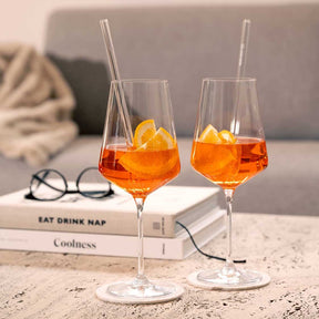 Zwei Weißweingläser PUCCINI mit orangefarbenem Getränk und Glasstrohhalmen auf Marmortisch, daneben Bücher und Brille