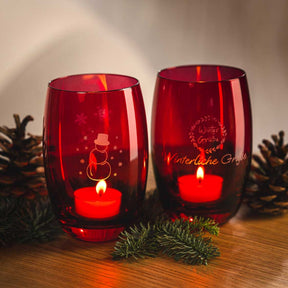Zwei rote Glastischlichter mit Gravur, brennenden Kerzen und Tannenzweigen auf Holztisch