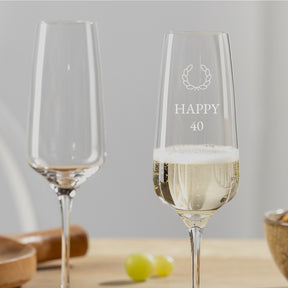 Zwei Sektgläser CESTI, eines mit Gravur "HAPPY 40" und Lorbeer-Motiv gefüllt mit Sekt, auf Holztisch
