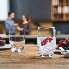Trinkglas CHEERS mit Eiswürfeln und roten Beeren auf Holztisch, im Hintergrund zwei Personen in Küche