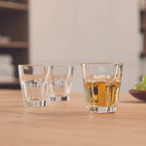 Stamper ROCK 50 ml mit Gravur auf Holztisch, gefüllt mit Whiskey, neben leeren Gläsern