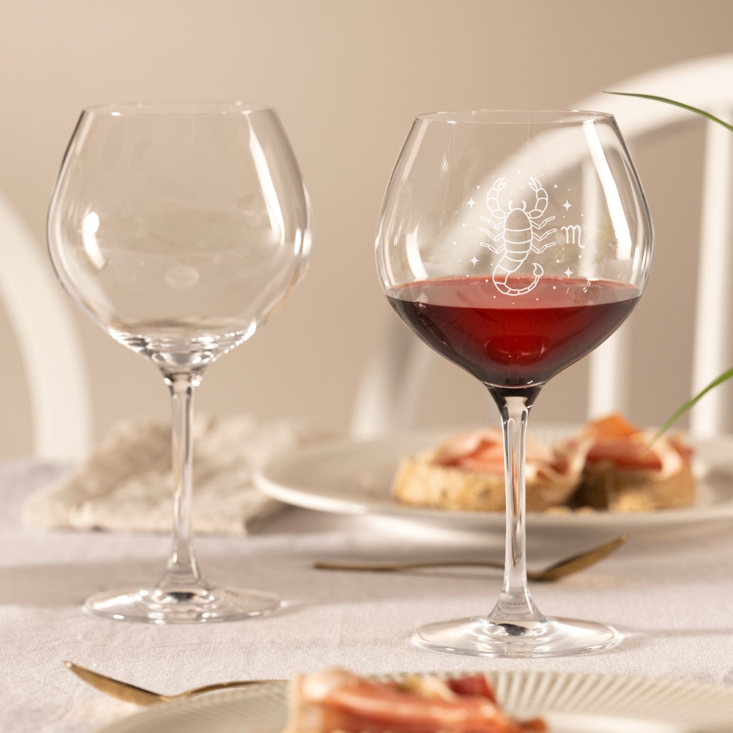 Burgunderglas CIAO+ mit Gravur, eines gefüllt mit Rotwein, auf gedecktem Tisch mit Gebäck