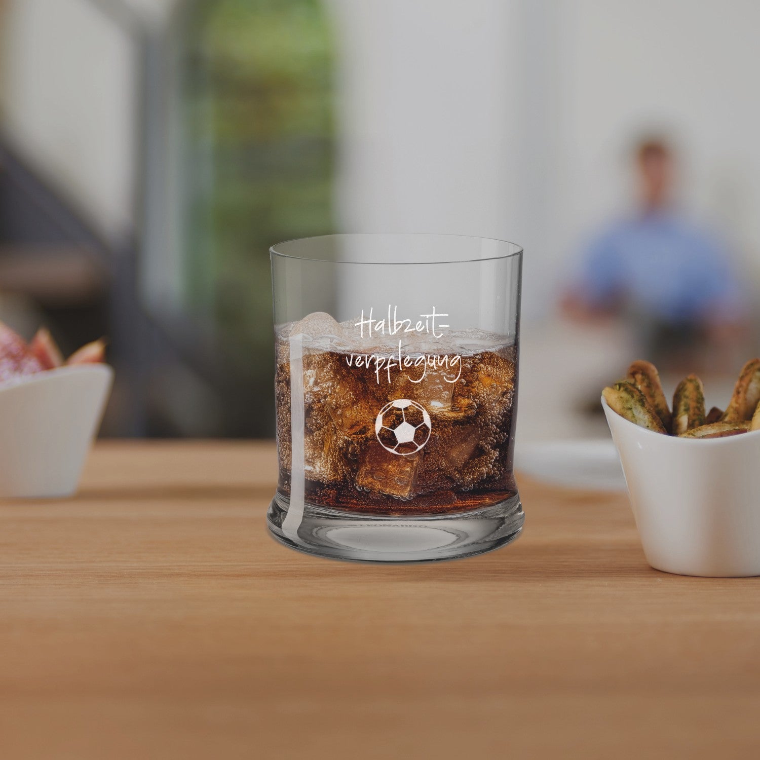 Trinkglas K18 mit Cola und Gravur auf Holztisch, umgeben von Snacks