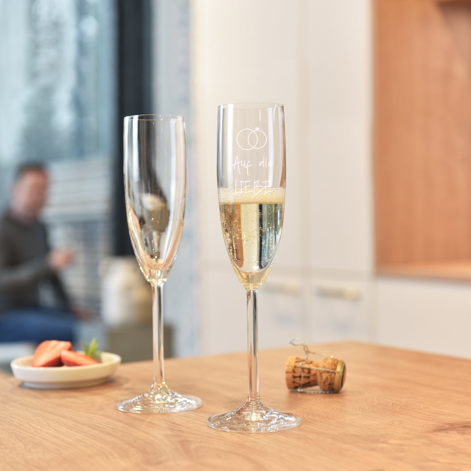 Zwei Sektgläser DAILY mit Gravur auf Holztisch, gefüllt mit Sekt, Büroscene im Hintergrund