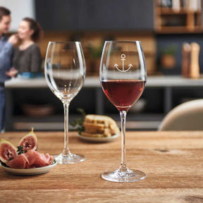Rotweinglas CHEERS mit Ankergravertung, gefüllt mit Rotwein, neben leerem Glas auf Holztisch in Wohnküche