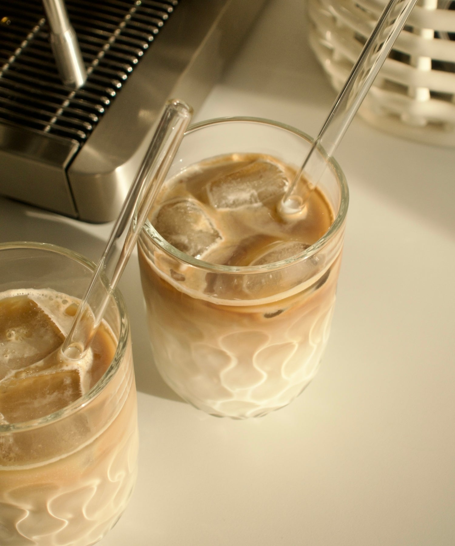 Zwei Gläser mit gemusterten Motiven gefüllt mit Eiskaffee und Strohhalmen, daneben eine Espressomaschine auf weißem Untergrund