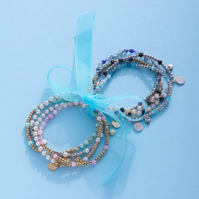 Zwei mehrfarbige Armbänder UNITA You & Me CIAO mit Perlen und Charms auf hellblauem Hintergrund