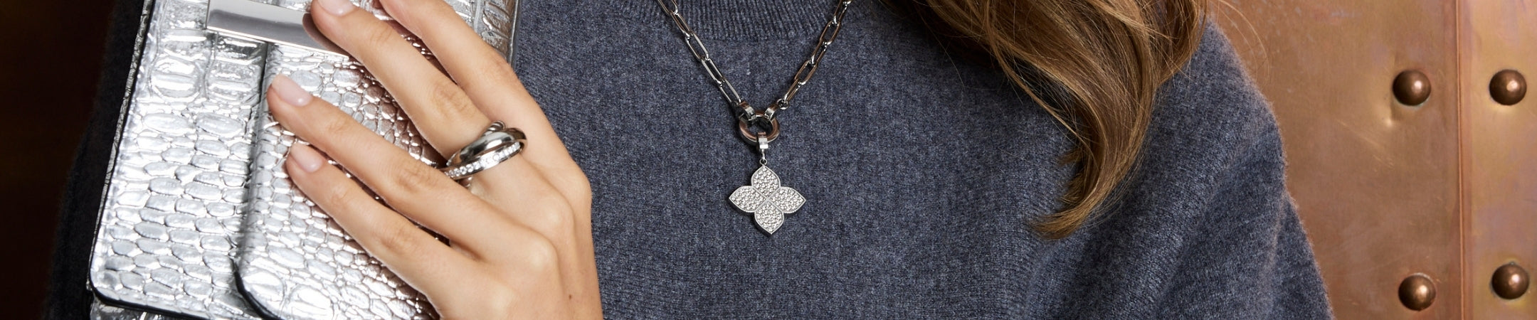 Frau trägt silbernen Anhänger mit Kleeblatt-Design an Halskette, dazu Ring und silberne Clutch, warme Farbstimmung